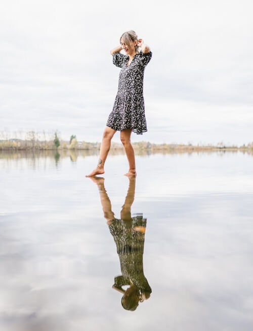Niki balanciert auf dem Wasser eines Sees, sie trägt ein schwarzes Kleid mit weißen Punkten und spiegelt sich in der glatten Oberfläche des Sees.